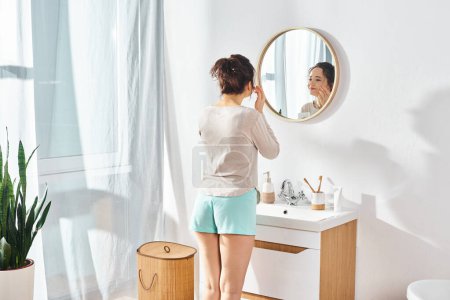 A brunette woman stands in front of a mirror in a modern bathroom, applying cream on face