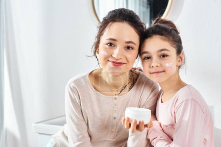 A brunette woman holds a cup of cream next to her preteen daughter in a modern bathroom, enjoying a beauty and hygiene routine.