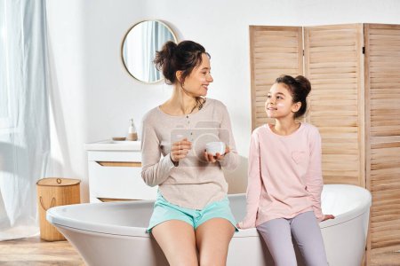 A brunette woman and her preteen daughter relax in a bathtub in a modern bathroom, enjoying a beauty and hygiene routine.