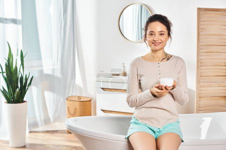 A stylish brunette woman holding a cream jar in her hand, sitting on bathtub