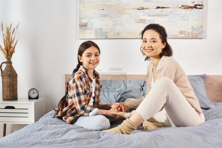 Zwei Frauen, Mutter und Tochter, sitzen auf einem Bett und lächeln freundlich in die Kamera in einer gemütlichen häuslichen Umgebung..