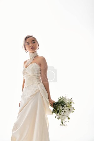 Photo for Attractive young asian woman in elegant white dress looking to side with bouquet of flowers - Royalty Free Image
