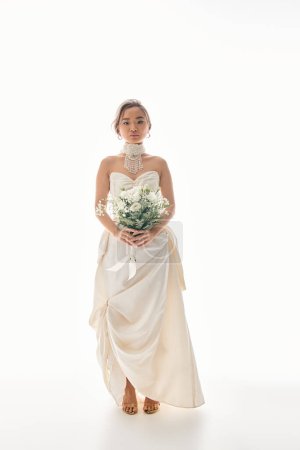 graceful asian bride in elegant dress holding to white flowers bouquet and looking to camera