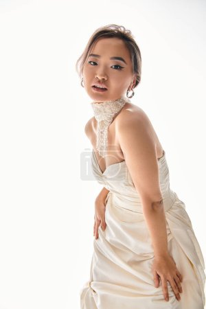 atractiva mujer asiática en elegante vestido blanco inclinado hacia adelante contra fondo claro