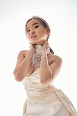Téléchargez les photos : Élégante asiatique jeune femme en robe blanche regarder caméra et étreindre cou avec les mains sur fond clair - en image libre de droit