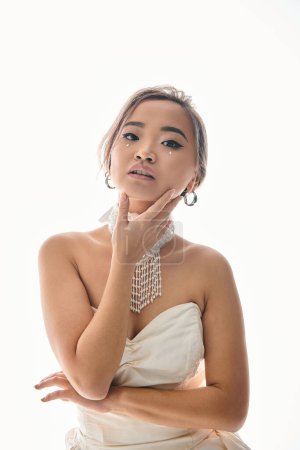 Téléchargez les photos : Élégante jeune mariée asiatique en collier blanc délicat toucher au visage avec la main sur fond clair - en image libre de droit