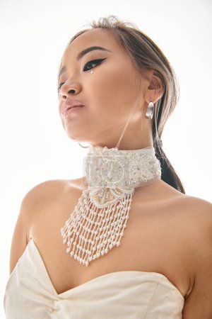 Téléchargez les photos : Séduisante asiatique jeune femme en blanc grâce collier avec élégant maquillage sur fond clair - en image libre de droit