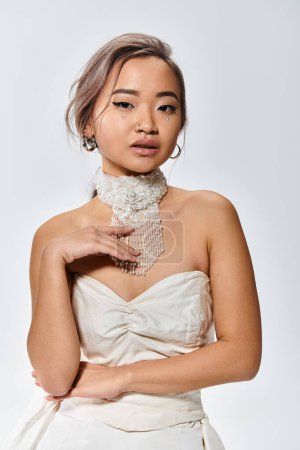 Téléchargez les photos : Élégante mariée asiatique délicate toucher à collier blanc et regardant à la caméra sur fond clair - en image libre de droit