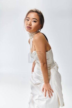 elegante novia asiática en elegante vestido blanco inclinado hacia adelante contra fondo claro