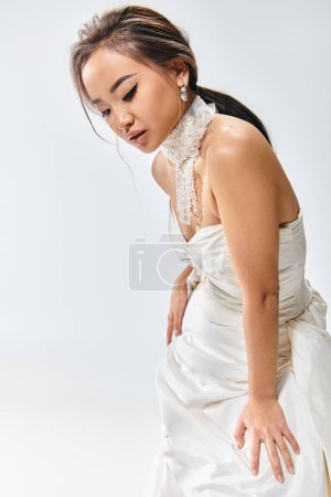 Foto de Encantadora chica asiática en elegante vestido blanco inclinado hacia adelante y mirando hacia abajo sobre fondo claro - Imagen libre de derechos