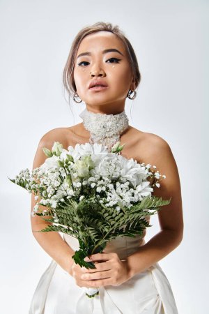 Téléchargez les photos : Gracieuse asiatique jeune mariée en blanc élégant tenue avec des fleurs bouquet sur fond clair - en image libre de droit