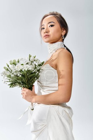 attractive asian bride in elegant outfit with white flowers bouquet looking sideways at camera