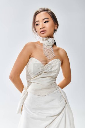 elegante mujer joven en vestido elegante blanco posando con las manos detrás de la espalda sobre fondo claro