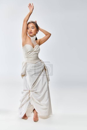 Photo for Alluring asian bride in white grace dress posing with hands and looking to side on light background - Royalty Free Image