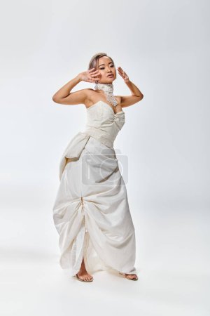 Foto de Encantadora novia asiática en blanco vestido de gracia posando con las manos cerca de la cara sobre fondo claro - Imagen libre de derechos