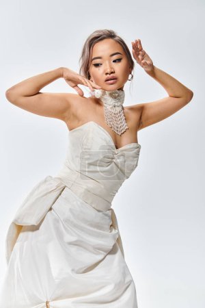 elegante asiático mujer en blanco gracia vestido posando con las manos cerca de cara y mirando hacia abajo