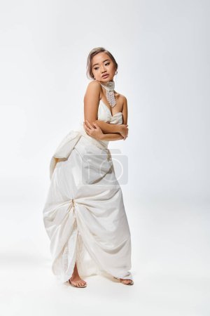 graceful asian young woman in white elegant dress hug herself and look to down on light background