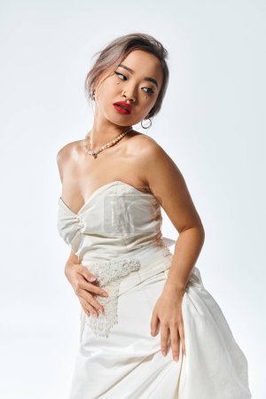 attractive asian bride with red lipstick and pearl necklace looking behind back on white background