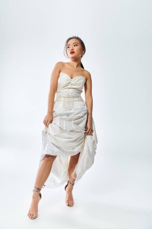 Foto de Atractiva mujer asiática en blanco elegante traje con labios rojos levantó su vestido y miró a un lado - Imagen libre de derechos