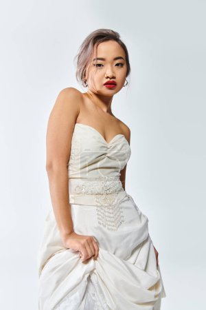 selbstbewusste asiatische Braut im weißen Anmut-Outfit mit roten Lippen hob ihr Kleid auf weißem Hintergrund
