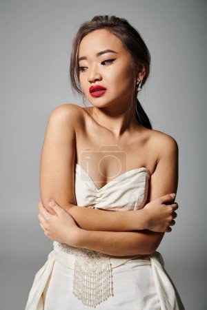 alluring asian young girl with pearl necklace and red lipstick hug herself on grey background