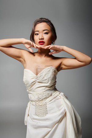 Téléchargez les photos : Séduisante asiatique mariée dans son 20s posant avec les mains dans le visage sur fond gris - en image libre de droit