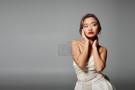 Téléchargez les photos : Attrayant asiatique jeune femme avec rouge lèvres regardant sur le côté et étreignant cou avec les mains - en image libre de droit