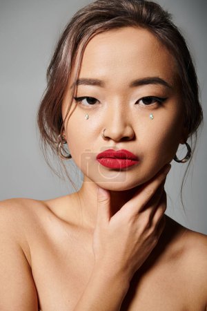 Téléchargez les photos : Portrait de la jeune femme asiatique élégante avec des lèvres rouges couvrant au cou avec la main sur fond gris - en image libre de droit