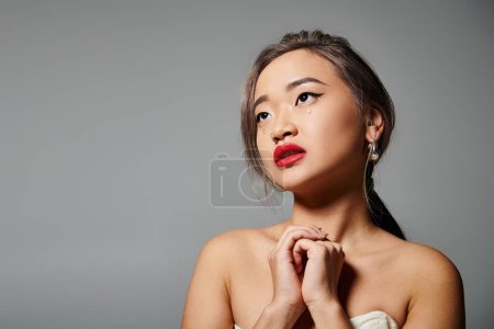 Photo for Attractive asian woman with red lips folding hands and looking to up against grey background - Royalty Free Image