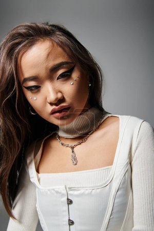 Charmante asiatische Frau mit gewagtem Make-up und Halskette lehnt sich nach vorne auf grauem Hintergrund