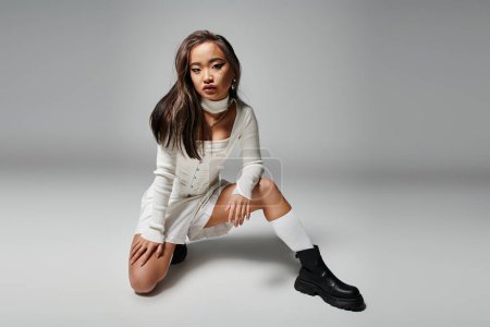 Wagemutiges asiatisches junges Mädchen in stylischem Outfit hockte sich vor grauem Hintergrund