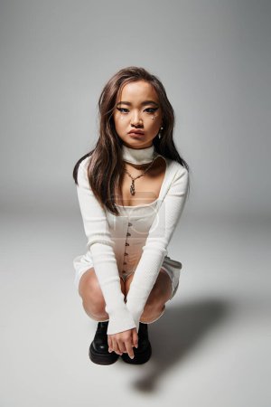hübsche asiatische junge Mädchen in weißen stilvollen Outfit hockte sich vor grauem Hintergrund