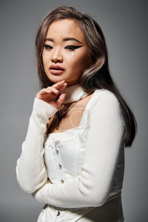 hermosa asiático joven mujer con outrageous maquillaje delicado tocar a barbilla en gris fondo