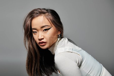 bastante asiático mujer en su 20s con pesado maquillaje de lado inclinado hacia adelante en gris fondo