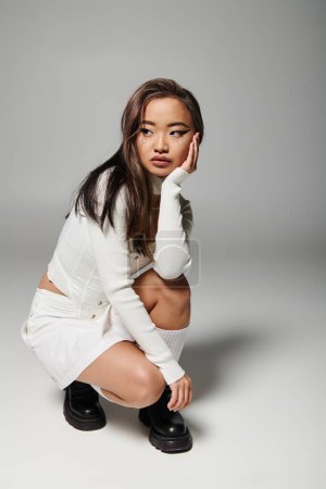 attractive asian woman in her 20s with heavy makeup sideways crouched down on grey background