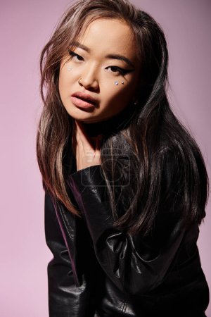 portrait of pretty asian woman in black leather outfit holding to neck with hand on lilac background