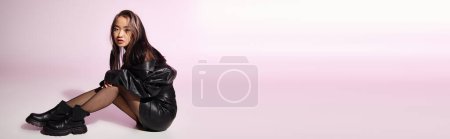 Téléchargez les photos : Bannière de femme asiatique en tenue en cuir noir avec un maquillage lourd assis latéralement sur fond lilas - en image libre de droit