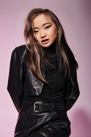 Photo for Pretty asian woman in black leather outfit with heavy makeup look to camera on lilac background - Royalty Free Image