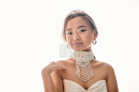Photo for Alluring asian young bride with grace necklace in white elegant dress touching to shoulder - Royalty Free Image