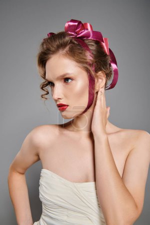 Une jeune femme respire la beauté classique dans une robe blanche avec un n?ud rose sur la tête, posant élégamment dans un décor de studio sur fond gris.