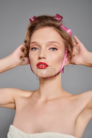 Une jeune femme à la beauté classique pose dans un studio, portant un arc rose sur la tête sur un fond gris.