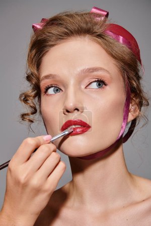 A young woman with a pink bow in her hair is carefully applying lipstick to her lips in a classic beauty pose on a grey background.