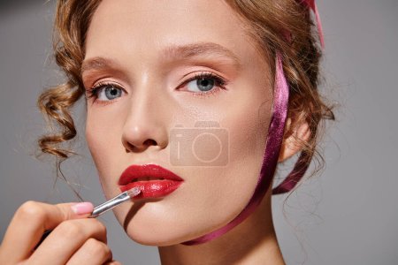 Photo for A young woman in a studio setting on a grey background, applying lipstick to enhance her lips with precision and care. - Royalty Free Image
