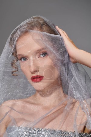 A young woman exudes classic beauty as she poses with a veil draped delicately over her head in a studio setting.