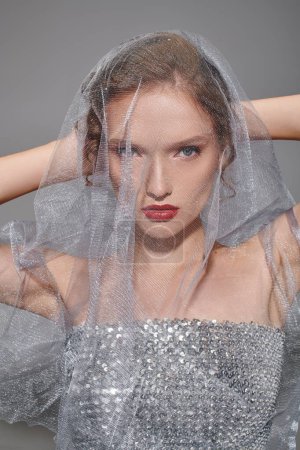 A young woman exudes classic beauty as she poses with a veil on her head in a studio setting against a grey backdrop.