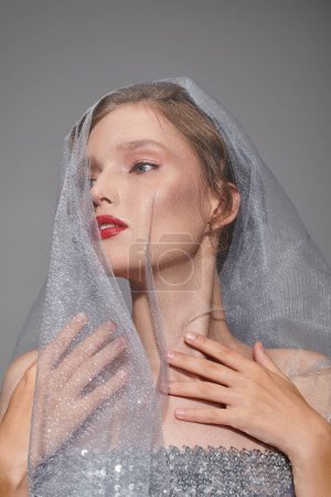 A young woman exudes classic beauty in a studio as she poses with a veil draped over her head.