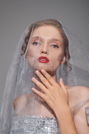 Eine junge Frau, die klassische Schönheit ausstrahlt, trägt einen Schleier und leuchtend roten Lippenstift in einem Studio vor grauem Hintergrund.
