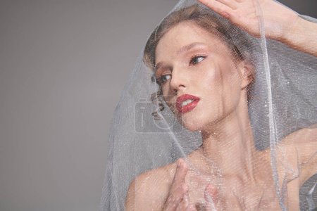 A young woman exudes classic beauty, posing gracefully with a veil on her head in a studio setting.