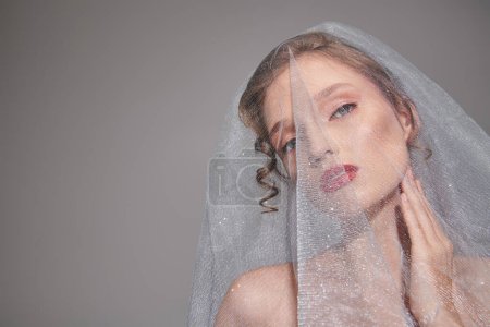 A young woman poses in a studio, wearing a veil on her head, exuding classic beauty and elegance.