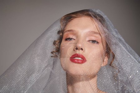 A young woman exudes classic beauty in a studio setting, wearing a veil and bold red lipstick.
